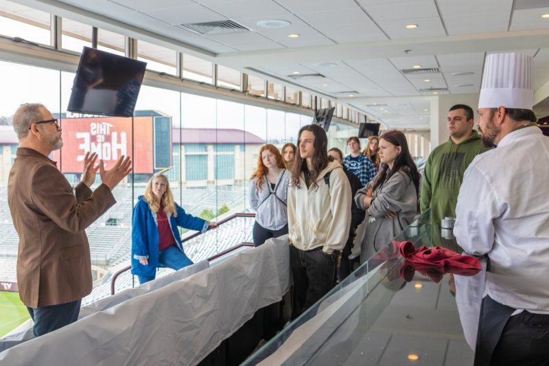 A group of high school students stand in a room overlooking the Virginia Tech football field listening to a man in a suit talk.